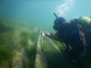 A diver in scuba gear is underwater and examining the lakebed using an object that looks like a digital camera. Below the diver, the lakebed is covered in sheets of hessian mats with some weeds poking through.